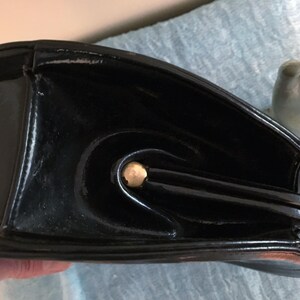 May include: Black patent leather handbag with a curved design and gold-tone hardware. The bag has a structured shape with a rounded base and a top handle. The glossy finish reflects light, highlighting the bag's details.