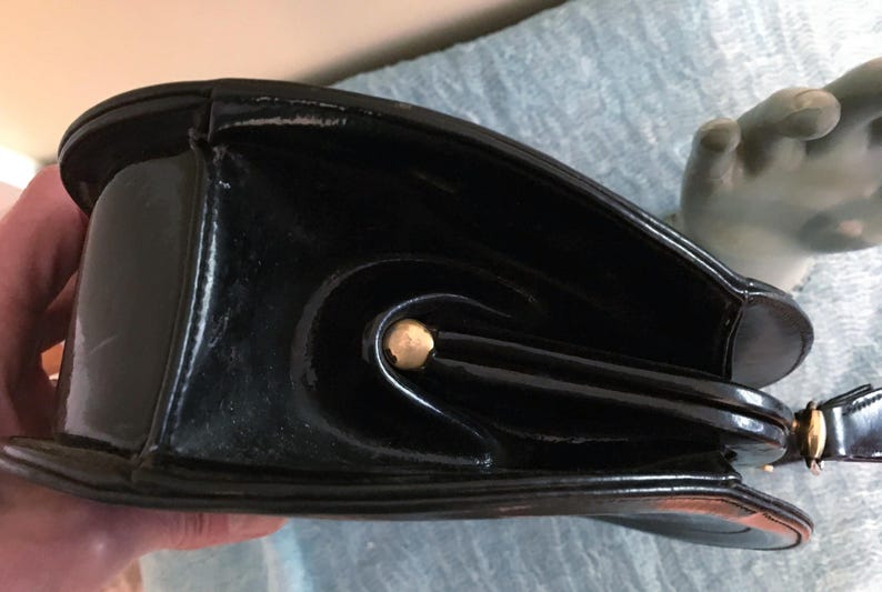 May include: Black patent leather handbag with a curved design and gold-tone hardware. The bag has a structured shape with a rounded base and a top handle. The glossy finish reflects light, highlighting the bag's details.
