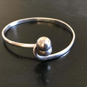 May include: A silver-toned bangle bracelet with a spherical accent. The bracelet features a simple, elegant design with a smooth, rounded band and a prominent, textured sphere. The bracelet is displayed on a dark surface.