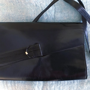 May include: Black leather crossbody bag with a rectangular shape. The bag features a long, adjustable strap and a decorative flap with a gold-toned accent. The bag's design is sleek and classic, suitable for various occasions.