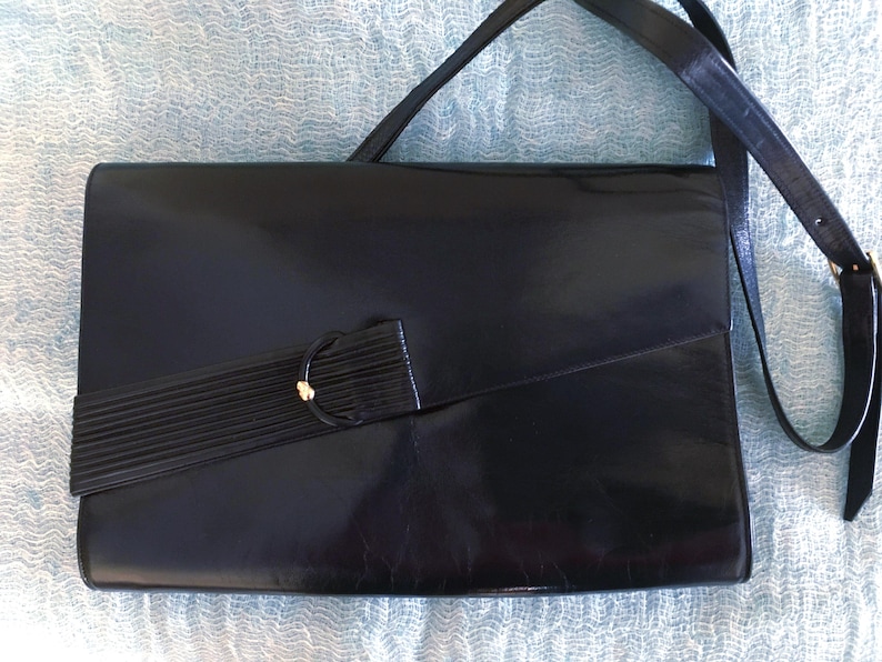 May include: Black leather crossbody bag with a rectangular shape. The bag features a long, adjustable strap and a decorative flap with a gold-toned accent. The bag's design is sleek and classic, suitable for various occasions.