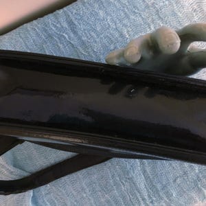 May include: A glossy black handbag with a shoulder strap. The rectangular bag has a shiny, patent leather-like finish. The bag is held by a hand with a light green color.