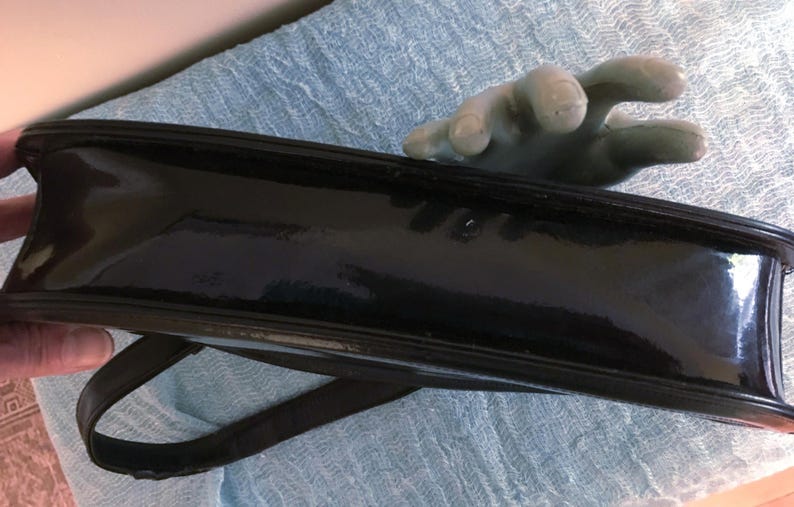 May include: A glossy black handbag with a shoulder strap. The rectangular bag has a shiny, patent leather-like finish. The bag is held by a hand with a light green color.