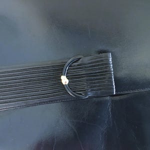 May include: Black leather clutch with a ridged flap and a black D-ring clasp with a gold accent. The clutch is made of a smooth, shiny leather material.
