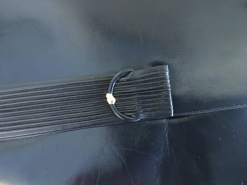May include: Black leather clutch with a ridged flap and a black D-ring clasp with a gold accent. The clutch is made of a smooth, shiny leather material.