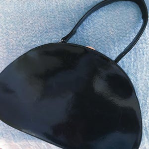 May include: A black, semi-circular handbag with a glossy finish. The bag has a single, curved handle and a smooth, reflective surface. The handbag is likely made of a patent leather or similar material.