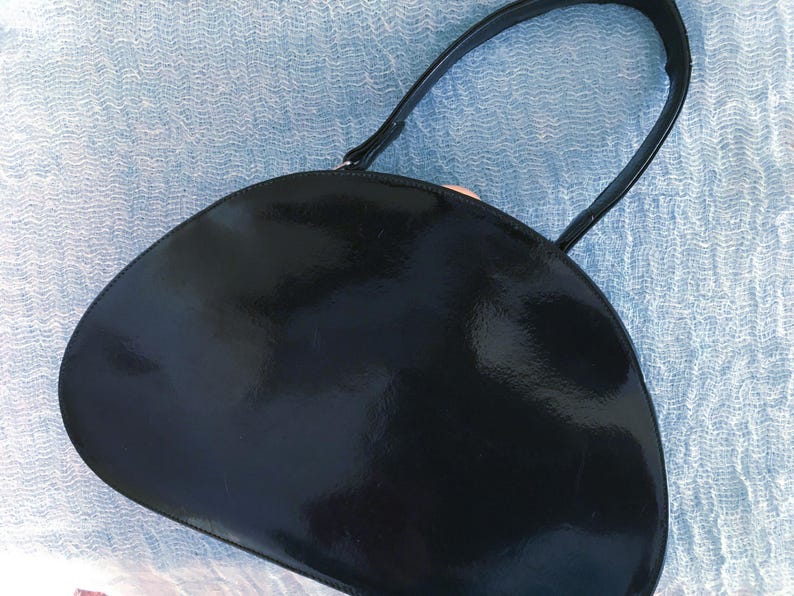 May include: A black, semi-circular handbag with a glossy finish. The bag has a single, curved handle and a smooth, reflective surface. The handbag is likely made of a patent leather or similar material.