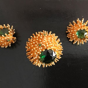Pu&ograve; includere: Un set di tre gioielli dorati con pietre preziose verdi. I pezzi hanno la forma di un'esplosione solare con una grande pietra preziosa verde al centro.