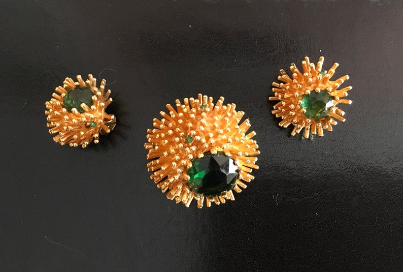 Pu&ograve; includere: Un set di tre gioielli dorati con pietre preziose verdi. I pezzi hanno la forma di un'esplosione solare con una grande pietra preziosa verde al centro.
