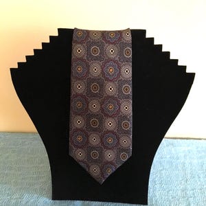 May include: A patterned necktie with a repeating floral design in shades of gray, blue, and red. The tie is displayed on a black velvet display stand. The tie's pattern is symmetrical and detailed.