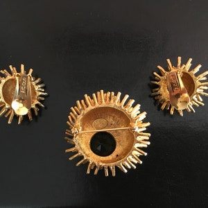 Pu&ograve; includere: Un set di tre gioielli dorati con un design a raggiera. Gli orecchini sono a clip e la spilla ha una pietra nera al centro. Il retro dei pezzi &egrave; contrassegnato con "Coro".