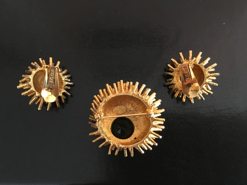 Pu&ograve; includere: Un set di tre gioielli dorati con un design a raggiera. Gli orecchini sono a clip e la spilla ha una pietra nera al centro. Il retro dei pezzi &egrave; contrassegnato con "Coro".