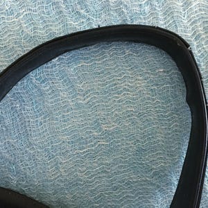 May include: Close-up of a black leather handbag handle. The handle is curved and shows signs of wear and tear. The background is a light blue, textured fabric.