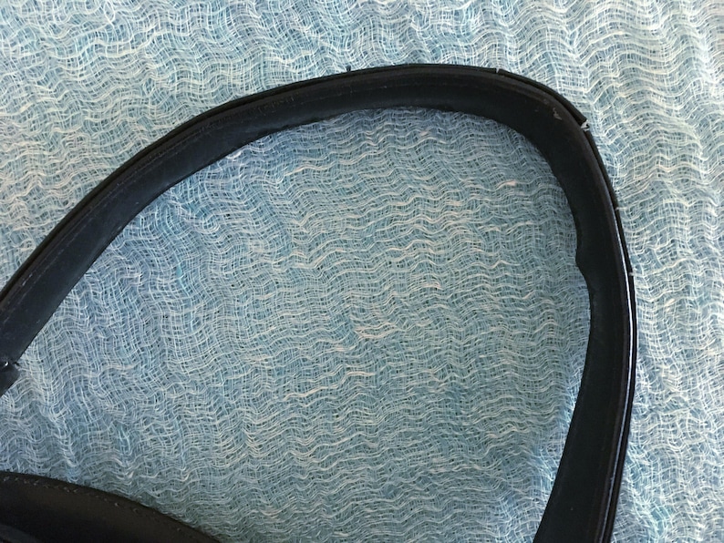 May include: Close-up of a black leather handbag handle. The handle is curved and shows signs of wear and tear. The background is a light blue, textured fabric.