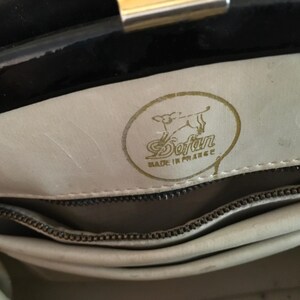 May include: Interior view of a black handbag with a gold clasp. The light-colored interior features a gold emblem with the text "Sofan" and "Made in France". A zipper is visible.
