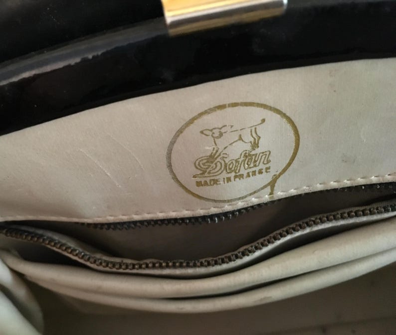 May include: Interior view of a black handbag with a gold clasp. The light-colored interior features a gold emblem with the text "Sofan" and "Made in France". A zipper is visible.