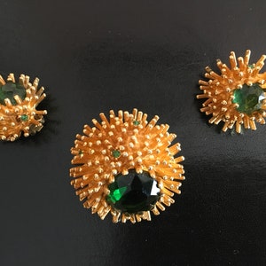 Pu&ograve; includere: Un set di tre gioielli dorati con pietre preziose verdi. I pezzi hanno la forma di un fiore spinoso o di uno scoppio di stelle.