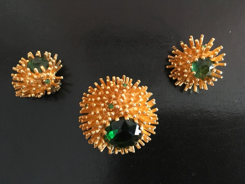 Pu&ograve; includere: Un set di tre gioielli dorati con pietre preziose verdi. I pezzi hanno la forma di un fiore spinoso o di uno scoppio di stelle.