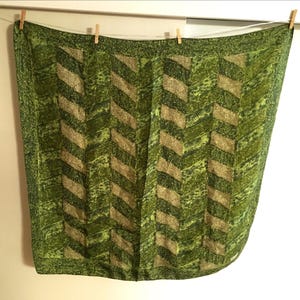 May include: A green and tan patterned scarf hanging from a clothesline. The scarf features a chevron design with alternating shades of green and tan. The edges of the scarf have a darker green border.