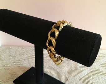 May include: A gold chain bracelet displayed on a black velvet jewelry stand. The bracelet is made of thick, chunky links.