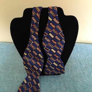 May include: Two blue bow ties with a repeating pattern of rectangles in red, beige, and navy blue. The bow ties are displayed on a black mannequin head against a light blue background. The bow ties are made of a patterned fabric.