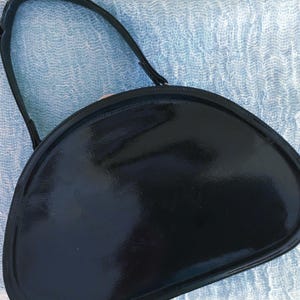 May include: A black, semi-circular handbag with a glossy finish. The bag has a single, thin handle and a rounded shape. The bag's surface reflects light, highlighting its smooth texture. The overall design is simple and elegant.