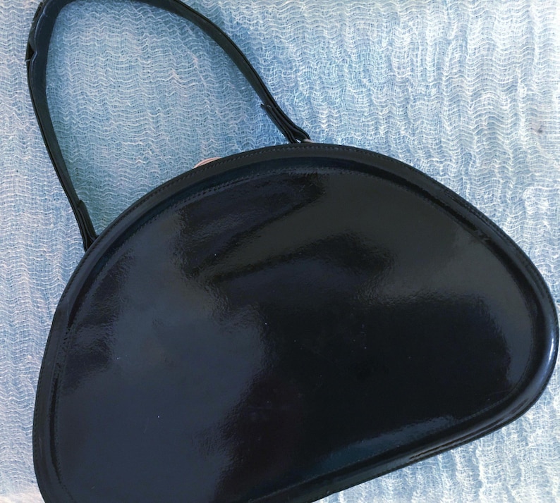 May include: A black, semi-circular handbag with a glossy finish. The bag has a single, thin handle and a rounded shape. The bag's surface reflects light, highlighting its smooth texture. The overall design is simple and elegant.