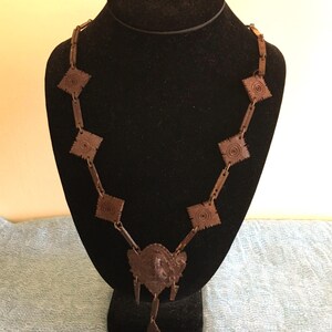 May include: A brown metal necklace displayed on a black mannequin. The necklace features square and rectangular links, with a central pendant and dangling diamond-shaped accents. The pendant has a face design.