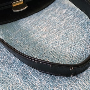 May include: Close-up of a black handbag with a silver-colored clasp and handle. The handle is made of black leather and shows signs of wear. The background is a light blue, textured fabric.