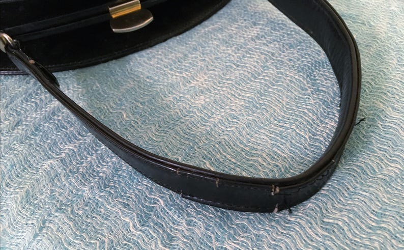 May include: Close-up of a black handbag with a silver-colored clasp and handle. The handle is made of black leather and shows signs of wear. The background is a light blue, textured fabric.