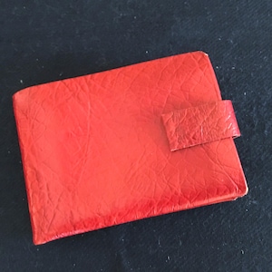 May include: A bright red wallet with a textured surface and a rectangular shape. The wallet has a small, matching red tab closure on the front. The wallet is closed.