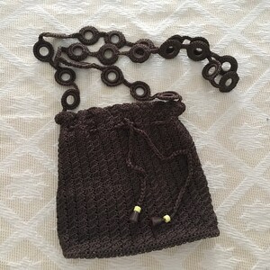 May include: A brown crocheted bag with a drawstring closure and a long strap with crocheted rings. The bag has two small yellow beads on the bottom.