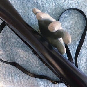 May include: A dark brown, glossy rifle with a black leather strap. The rifle is held by a gray hand-shaped object. The strap is attached to the rifle with a gold-colored buckle. The background is a light blue, textured fabric.