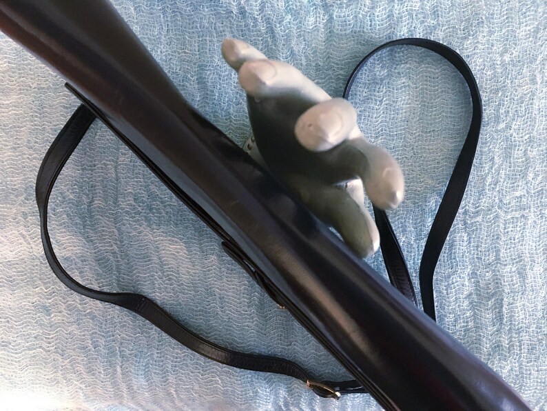 May include: A dark brown, glossy rifle with a black leather strap. The rifle is held by a gray hand-shaped object. The strap is attached to the rifle with a gold-colored buckle. The background is a light blue, textured fabric.