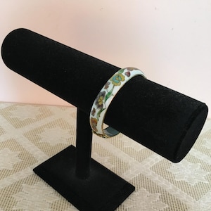 Op de afbeelding: Witte emaille armband met een bloemenmotief in tinten oranje, geel, groen en bruin. De armband ligt op een zwarte sieradenstandaard.