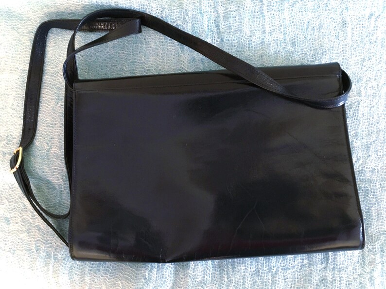 May include: A black leather crossbody bag with a rectangular shape. The bag has a long, thin strap and a gold-colored buckle. The leather appears shiny and smooth, with a subtle sheen.