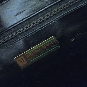 May include: Close-up of a black leather handbag with a zipper and a gold-toned rectangular label. The label reads "BRUNO MAGLI MADE IN ITALY BOLOGNA" with a stylized "M" logo.