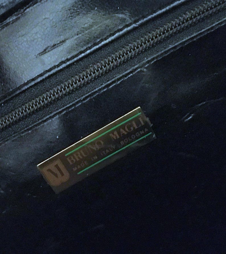 May include: Close-up of a black leather handbag with a zipper and a gold-toned rectangular label. The label reads "BRUNO MAGLI MADE IN ITALY BOLOGNA" with a stylized "M" logo.