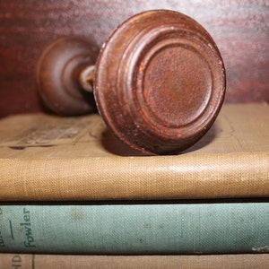 May include: A stack of three vintage books with brown, blue, and beige covers. The top book has the title "1000 Things Worth Knowing" and the author "Fowler". The bottom book has the title "Everyday Etiquette" and the author "Harland". The middle book has the title "Bobbs-Merrill" and the author "Sully". A wooden door knob sits on top of the books.