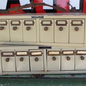May include: Two stacked metal drawer cabinets with labeled drawers. The top cabinet has six drawers and the bottom cabinet has eight drawers. Both cabinets are painted a light beige color.