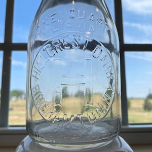 May include: A clear glass milk bottle with a white cap. The bottle is embossed with the text "One Quart Liquid", "Hillcrest Dairy", "Binghamton, N.Y.", and a large "H" in a circle.
