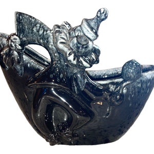 May include: A black and white ceramic planter shaped like a clown holding a ball. The planter has a glazed finish and is in the shape of a boat.