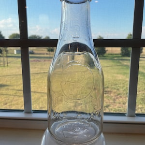 May include: A clear glass milk bottle with a rounded bottom and a narrow neck. The bottle is embossed with the text "ONE QUART" and a logo that appears to be a stylized letter "C".