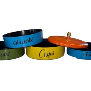 May include: Four round snack containers with lids. The containers are blue, green, yellow, and orange. The lids are black. The containers are labeled "Snacks", "Chips", "Nuts", and "Snacks".