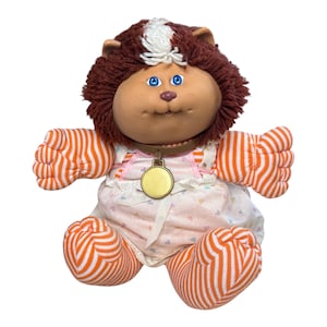May include: A vintage Cabbage Patch Kids doll with a brown, shaggy hairstyle and blue eyes. The doll wears a pink and white dress with a heart pattern and orange and white striped arms and legs. It has a gold-colored medallion around its neck.