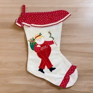 May include: A white Christmas stocking with a red and white ruffled top. The stocking features an embroidered image of a man in a red suit and a hat holding a stick. The man is wearing a red and white suit and a hat. The stocking has the words "Merry Christmas" embroidered on it.