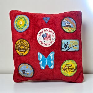 May include: Red square pillow adorned with various embroidered patches. Patches include a butterfly, the American flag with "Keep America Beautiful", and other patches with text like "Cookie Olympics 1980" and "Camp Cibola 1978".