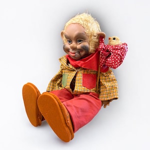 May include: A vintage doll with a painted face, blonde hair, and large ears. It wears a plaid shirt, red pants, and orange shoes. A red scarf and a bird with a polka-dot bag are also featured. The doll is a collectible item.