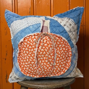 May include: A square quilted throw pillow featuring a patchwork design of blue and white patterned fabrics. Centered on the pillow is an orange pumpkin applique with a white floral print, a plaid stem, and a decorative jute twine bow tied at the top.