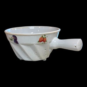 May include: White ceramic bowl with a handle, featuring a spiral design and small holes. The bowl is decorated with painted strawberries and purple flowers. The handle has a small hole. The bowl is on a black background.